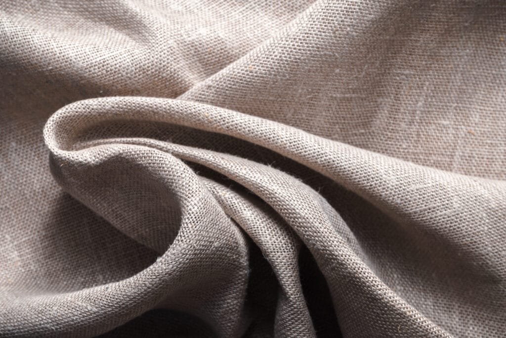 Close-up of soft, natural linen fabric with a plain weave and subtle texture, showing gentle folds and a warm beige-gray color.