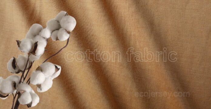 Close-up of warm tan cotton fabric with a subtle weave, accented by a cluster of white cotton bolls on the left.