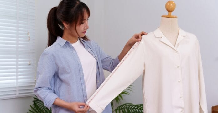 A woman in a light blue shirt and white t-shirt is measuring the sleeve of a white button-up blouse on a dress form using a measuring tape.