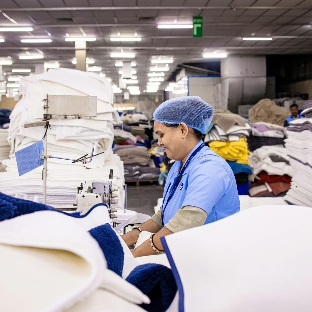 A factory worker wearing a blue uniform and a protective blue hairnet operates a sewing or binding machine in a large textile factory. Stacks of folded fabrics and blankets surround her, with bright overhead lights and a busy, organized workspace.

