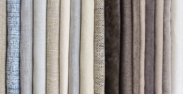 A row of rolled fabric bolts in various neutral linen tones, ranging from light beige to taupe and gray, showing different textures and weave patterns.