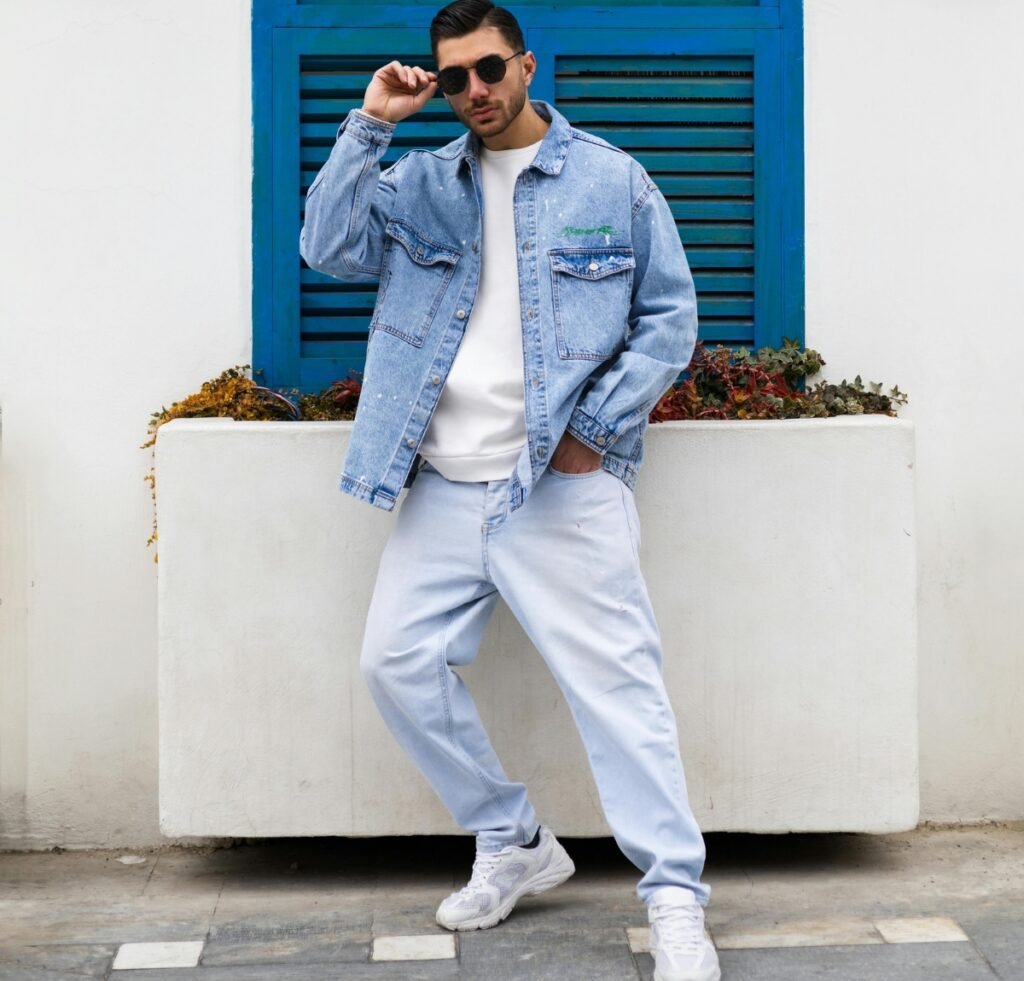 A stylish man wearing a light blue denim bomber jacket over a white T-shirt and matching light-wuel denim jeans, posing confidently against a white wall with a bright turquoise window behind him; he wears sunglasses and white sneakers.