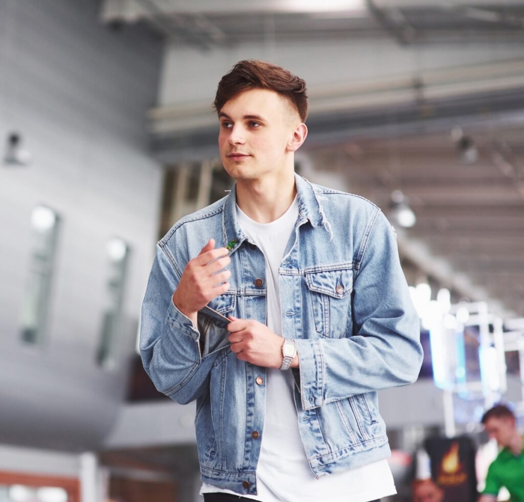 A young man wearing a light-wash denim bomber jacket over a white T‑shirt, in an indoor modern space. He adjusts the jacket’s collar with one hand, and a silver wristwatch is visible on his other wrist. The background features blurred architectural details and soft lighting.
