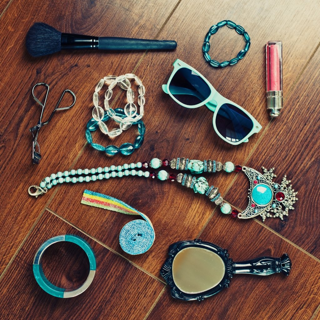 Assorted fashion accessories scattered on a wooden floor, including a makeup brush, sunglasses, beaded bracelets, a tiara, scissors, a lipstick, a blue velvet ribbon, and a decorative mirror.