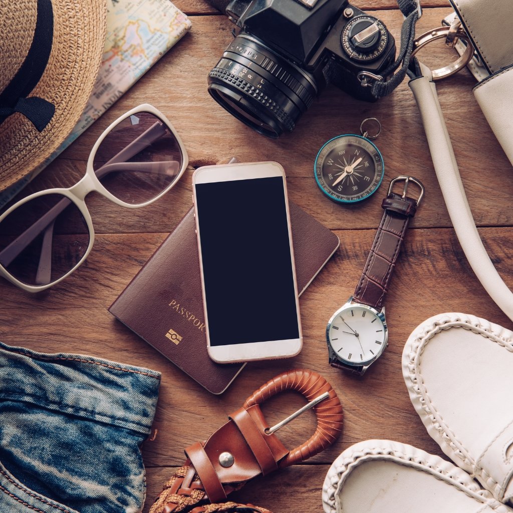 A flat lay image of travel accessories on a wooden surface, including a brown passport, smartphone, watch, sunglasses, camera, compass, belt, denim fabric, and white loafers; velour/wool fabric texture visible in the textile elements.