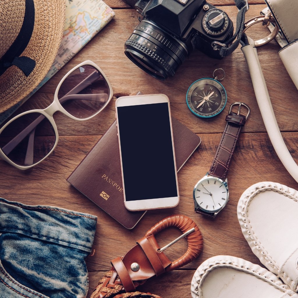 A flat lay of travel essentials on a wooden surface, including a camera, passport, smartphone, sunglasses, compass, wristwatch, belt, belt buckle, denim fabric, white loafers, and a woven hat; the items rest around angora wool fabric accents.