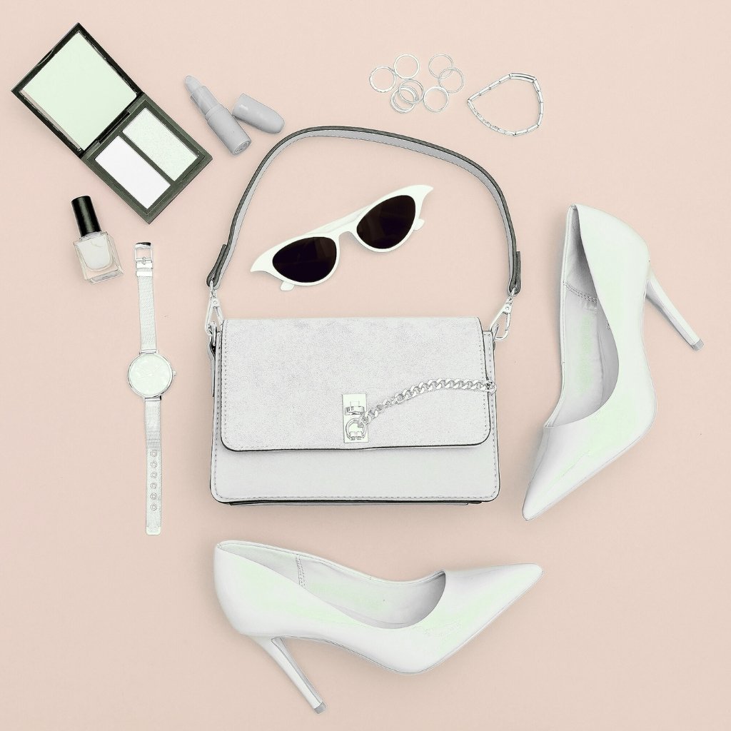 A flat lay of a stylish outfit and accessories on a pale pink background, including a white handbag with chain, white high-heeled shoes, white sunglasses, a silver watch, nail polish, makeup kit, rings, bracelet, and a few other small items.