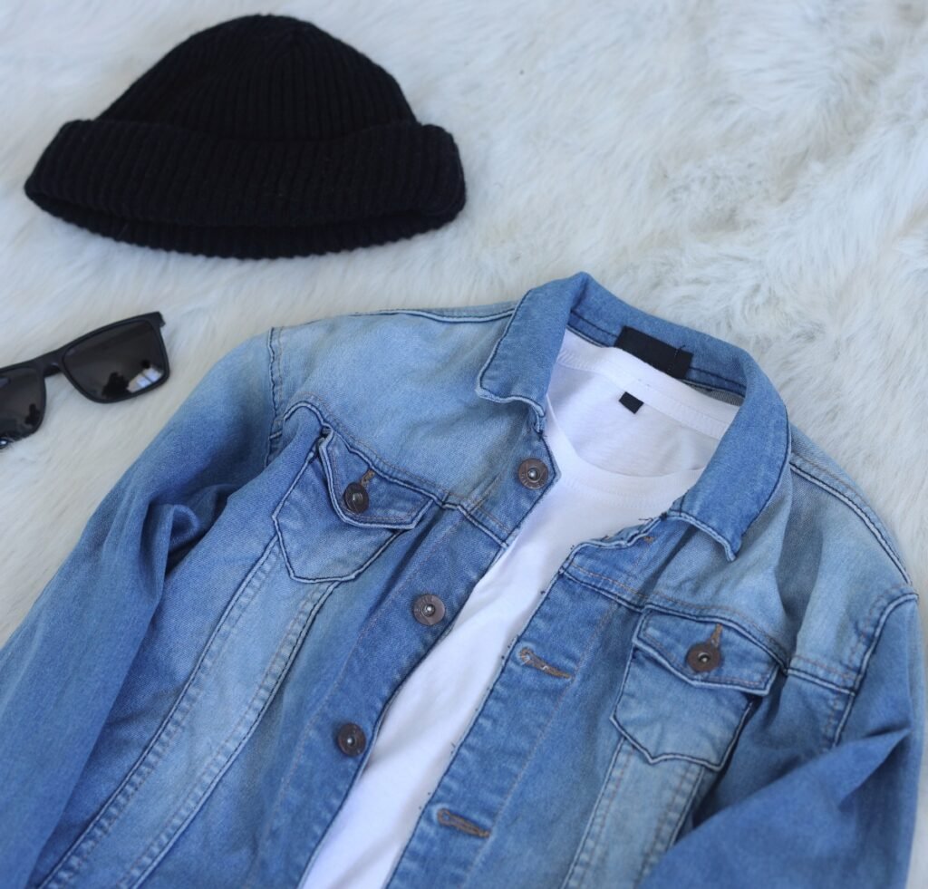 A light blue denim bomber jacket laid flat on a soft, white faux fur surface, with a white t-shirt underneath. A black knit beanie and a pair of black sunglasses sit nearby.