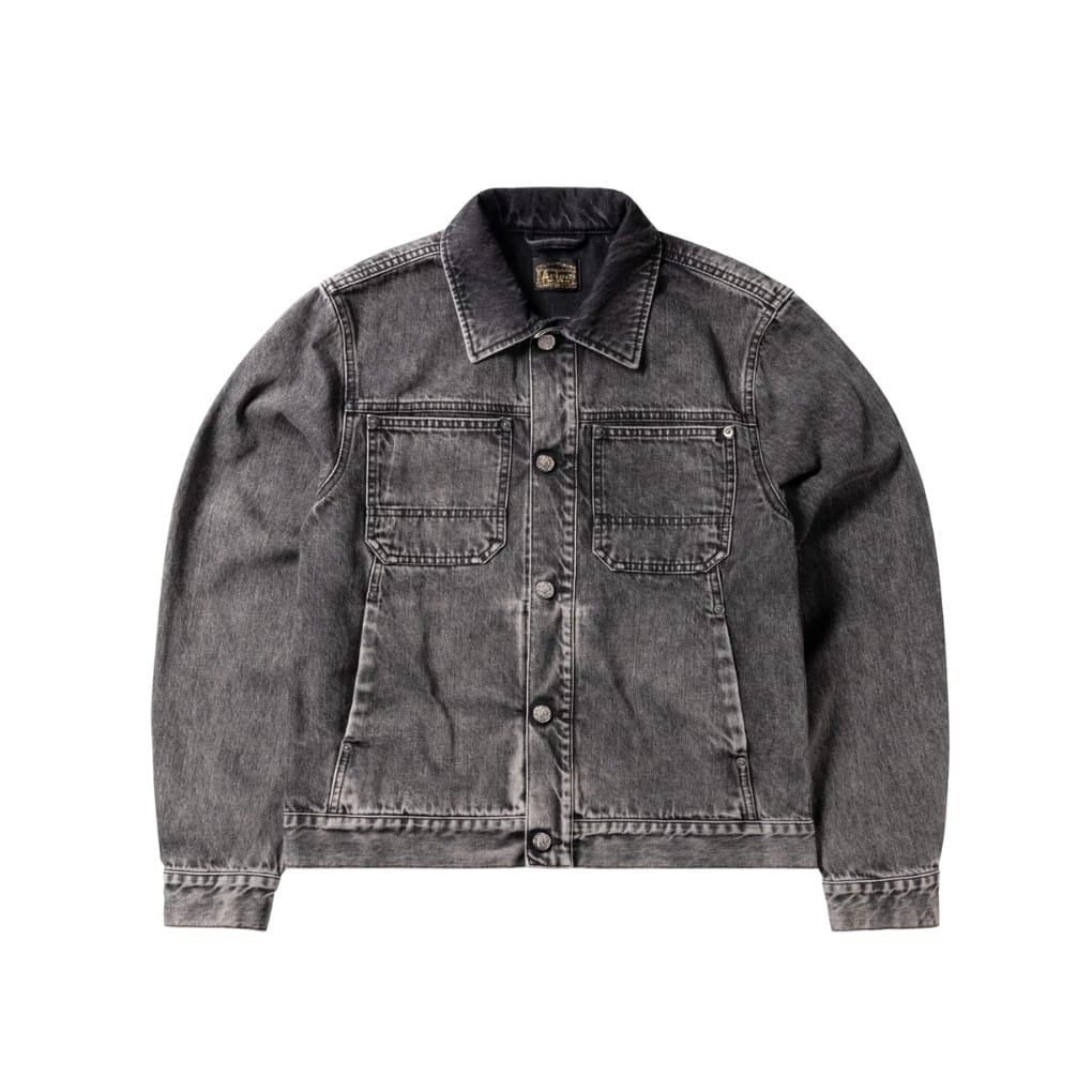 A grey-black washed denim jacket with a faded finish, two chest pockets, metal buttons, and a black collarless look, displayed against a white background.