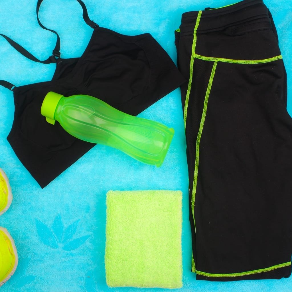 A flat lay of athletic workout gear on a bright blue background: black sports bra and black leggings with neon green stitching, uses of polyester fabric a neon green water bottle, a lime green washcloth, and part of yellow sneakers visible. uses of polyester fabric