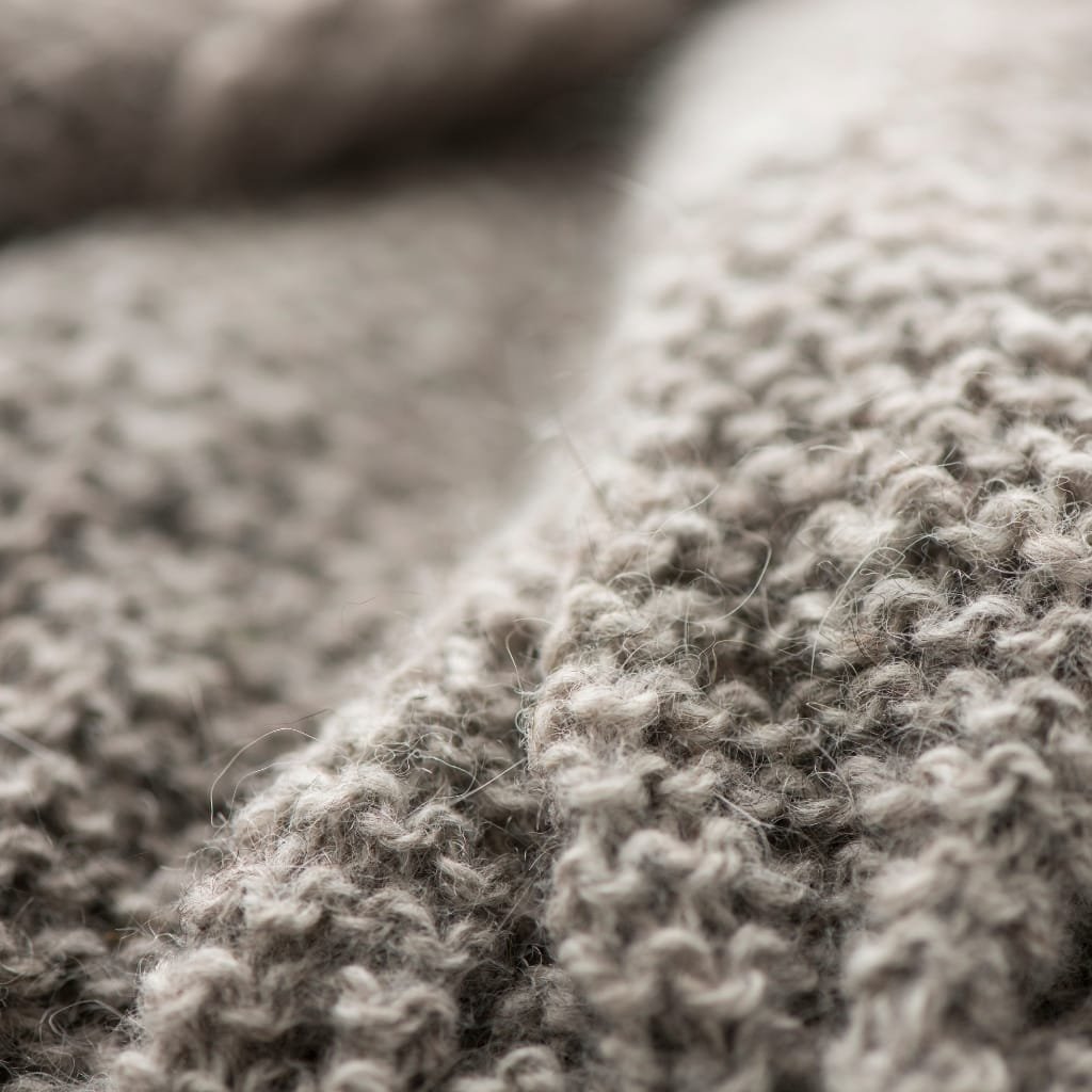 A close-up macro shot of soft, chunky gray wool fabric. The textured stitches show a woven knit pattern with fuzzy fibers and natural variation in shade.