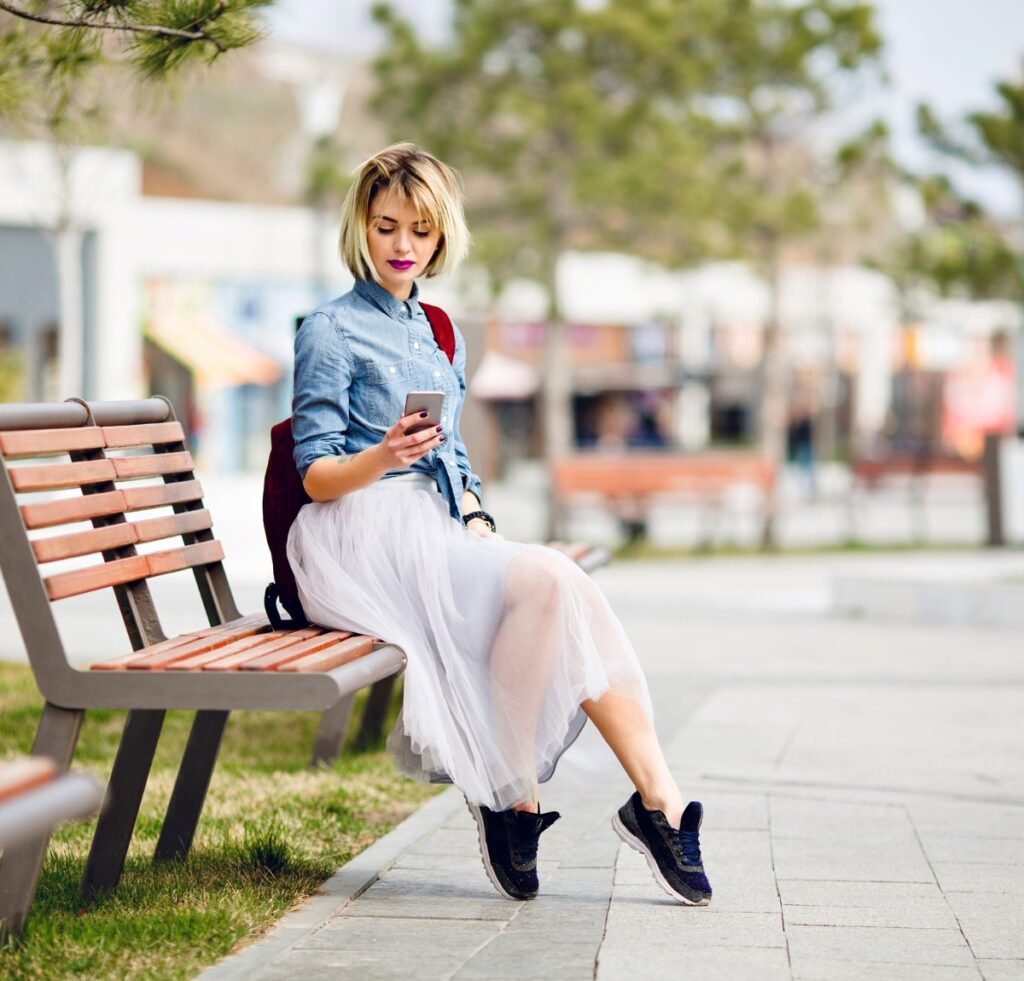 A stylish young woman sits on a park bench outdoors, looking at her smartphone. She wears a light denim shirt jacket over a pale tulle skirt, black sneakers, and carries a burgundy backpack. The scene is bright and blurred in the background with trees and benches.