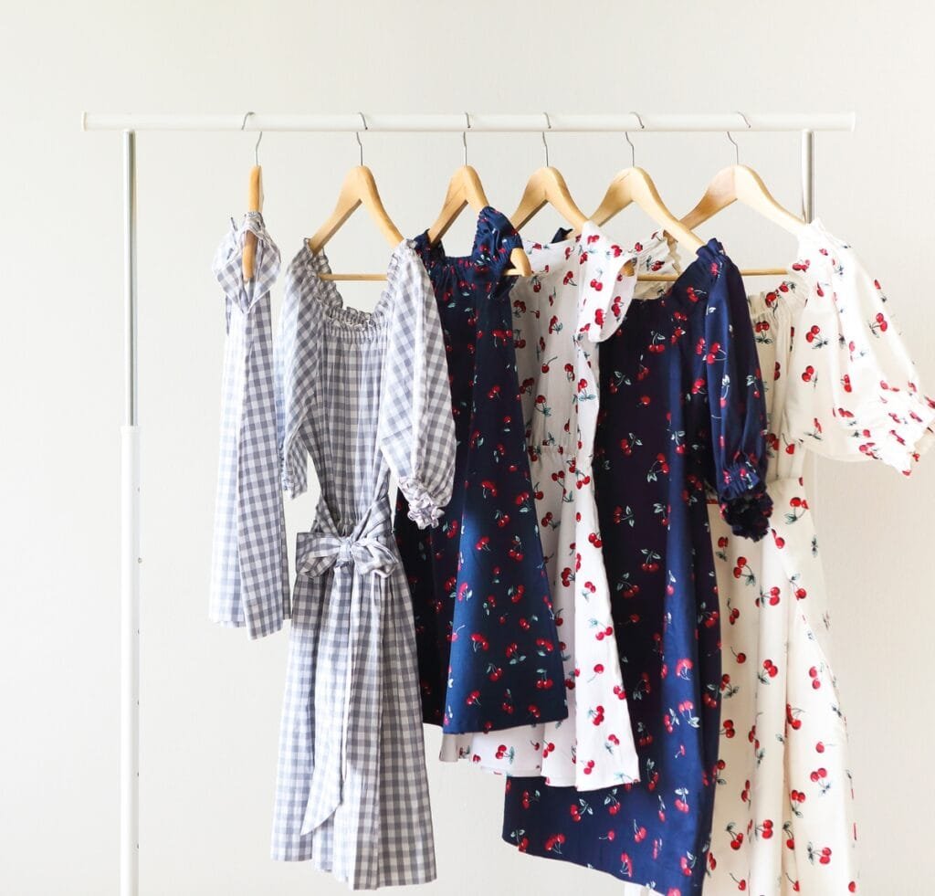 "A row of women's dresses and blouses hanging on wooden and metal hangers on a white clothing rack. The garments feature light blue gingham, navy and white prints, floral patterns, and puff sleeves, showcasing a variety of woven fabrics and textures."