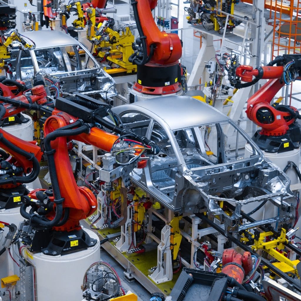 An advanced automotive manufacturing plant with multiple orange robotic arms assembling a silver car body. Robotic arms, conveyors, and support structures fill a busy, high-tech factory floor.