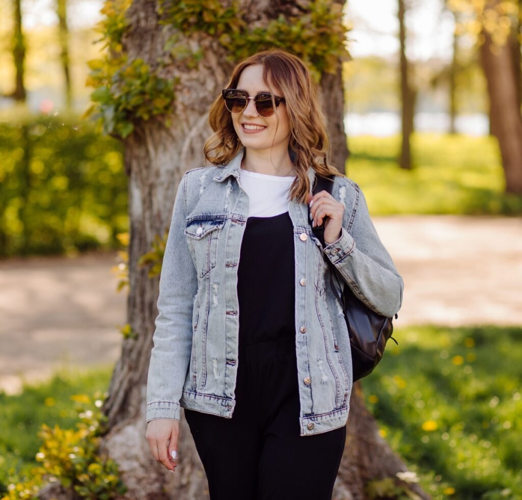 A smiling young woman wearing a light-wash denim bomber jacket over a black outfit with a white top, sunglasses, and a black backpack, standing outdoors near a tree in a sunlit park.