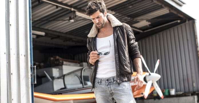 A man wearing a dark brown B-3 bomber jacket with a shearling collar, white graphic tee, and faded jeans stands near an open aircraft hangar. He holds sunglasses in one hand as he looks downward.