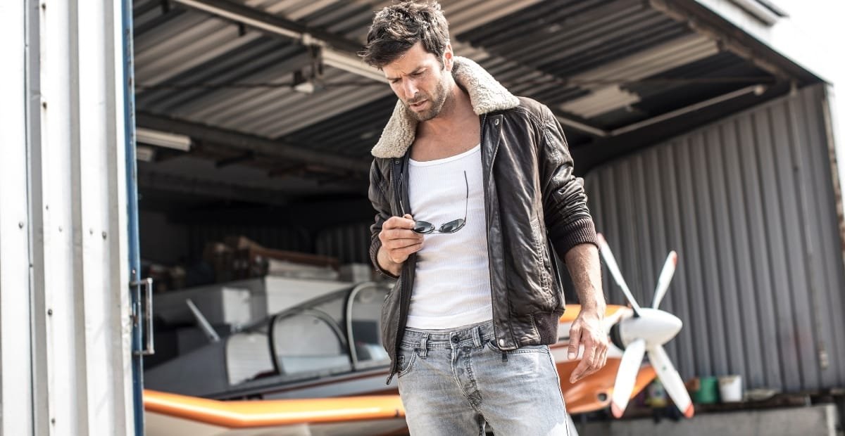 A man wearing a dark brown B-3 bomber jacket with a shearling collar, white graphic tee, and faded jeans stands near an open aircraft hangar. He holds sunglasses in one hand as he looks downward.