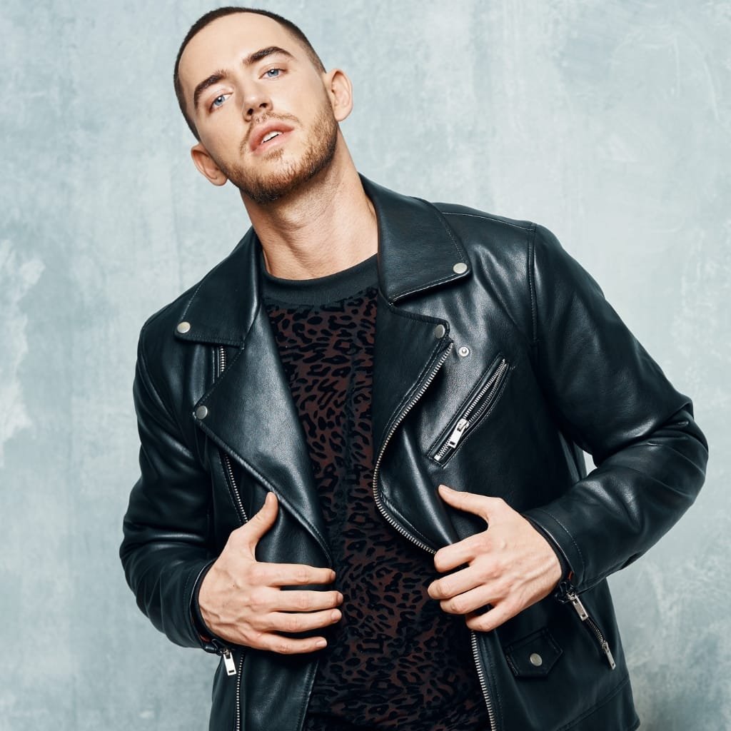 A man wearing a black leather biker jacket over a dark leopard print shirt, posing against a light gray textured wall. He gazes slightly upward with a relaxed demeanor, hands near his jacket, showcasing the zipper details