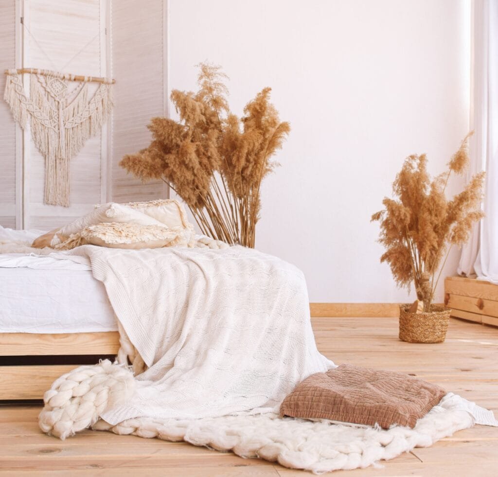 A cozy, neutral-bedroom scene featuring soft beige and cream Bird’s Eye linen fabrics. A bed is draped with a textured white-knit blanket and layered pillows. Ornamental dried pampas grass in woven baskets grouping beside the bed adds natural warmth to the light wood floor and white walls.
