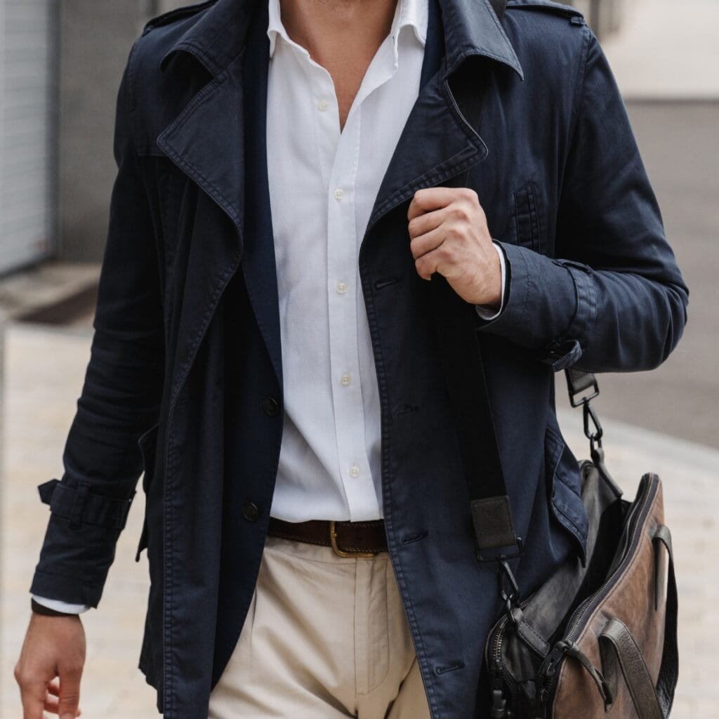A man wearing a navy blazer-style trench over a white button-down shirt, paired with light beige chinos. He carries a brown leather messenger bag with a detachable strap slung over his shoulder.
