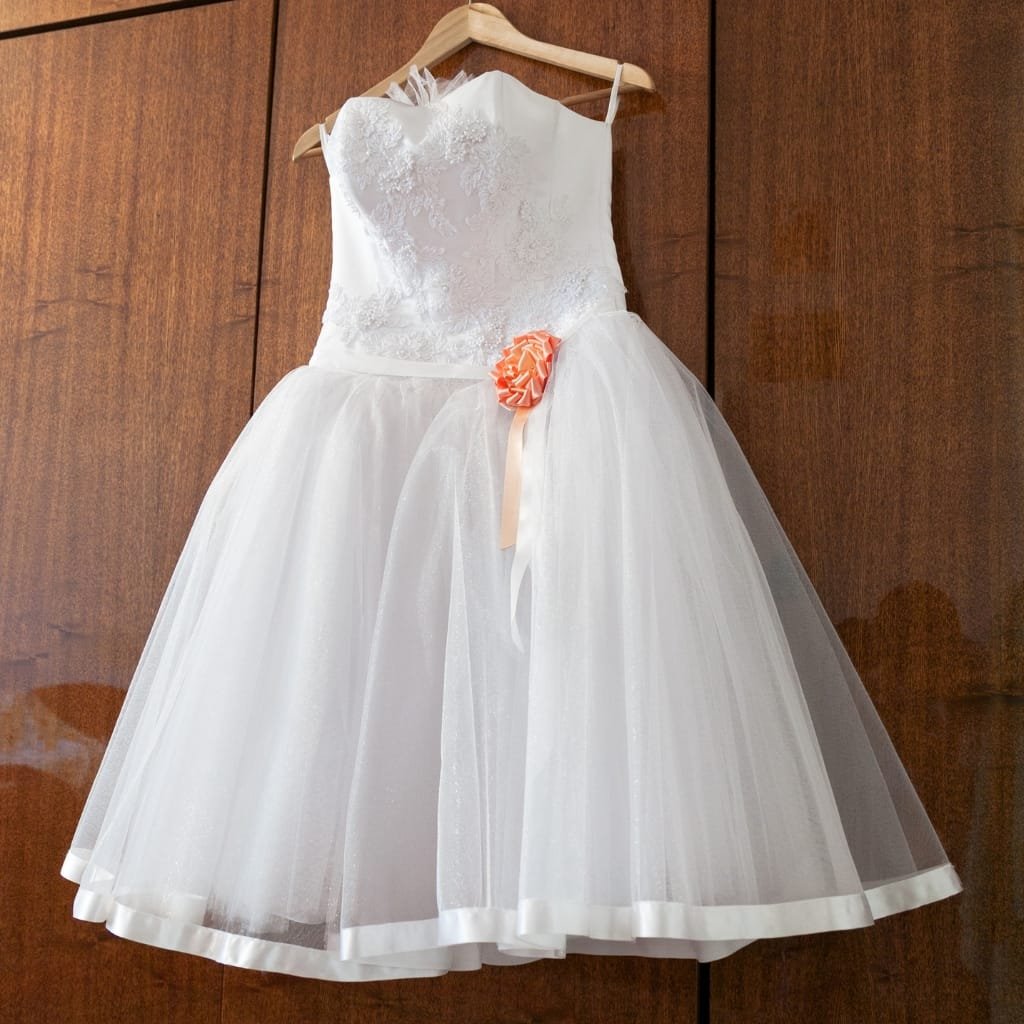 A white Broderie Anglaise cotton dress hanging on a wooden hanger; delicate floral embroidery on the bodice with a full tulle skirt and satin trim at the hem.

