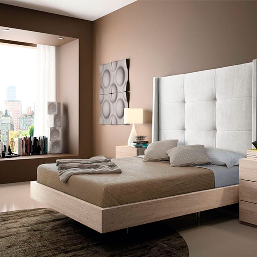 A modern bedroom with a light wood platform bed and a tall, upholstered headboard. A soft taupe bed cover, blue-grey sheets, and fluffy pillows sit on the bed. A nightstand with a lamp sits beside, and a large window with a city view is in the background. Neutral brown walls and a dark area rug complete the look.