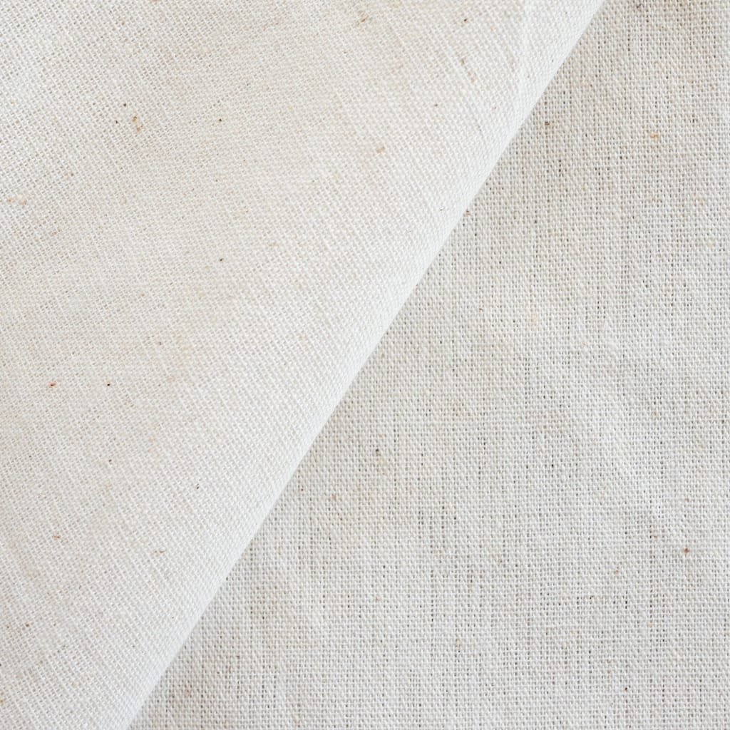 Close-up image of natural calico cotton fabric with a light beige weave, showing tiny specks and a folded corner.