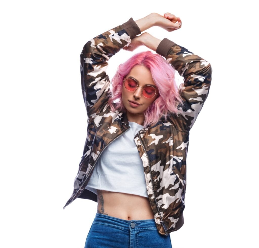 "Person wearing a camouflage bomber jacket with brown, tan, and white patterns, white cropped top, and blue jeans. The individual has pink hair and pink-tinted sunglasses, posed with arms raised over the head against a plain white background."