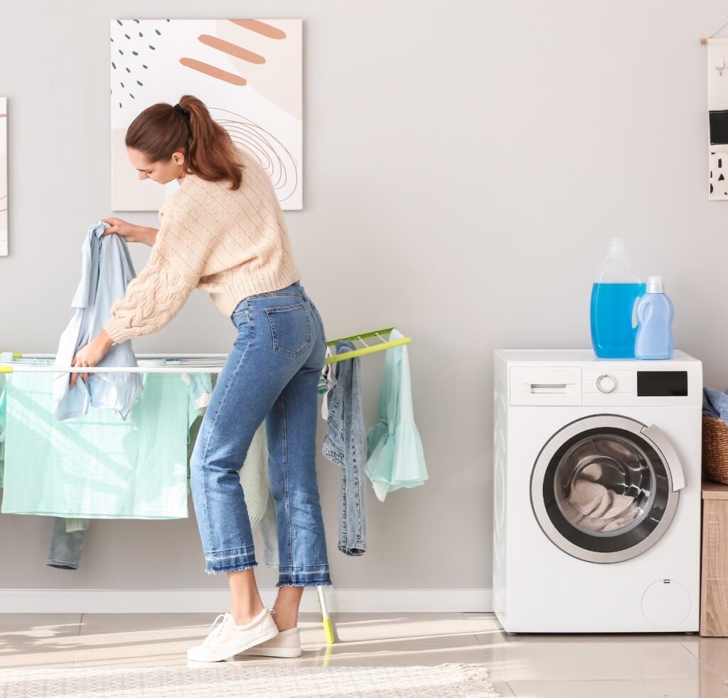 A woman drying laundry indoors: she stands by a drying rack, hanging light blue garments. A front-loading washing machine is nearby with laundry supplies on top; the room has soft neutral walls and decorative wall art.