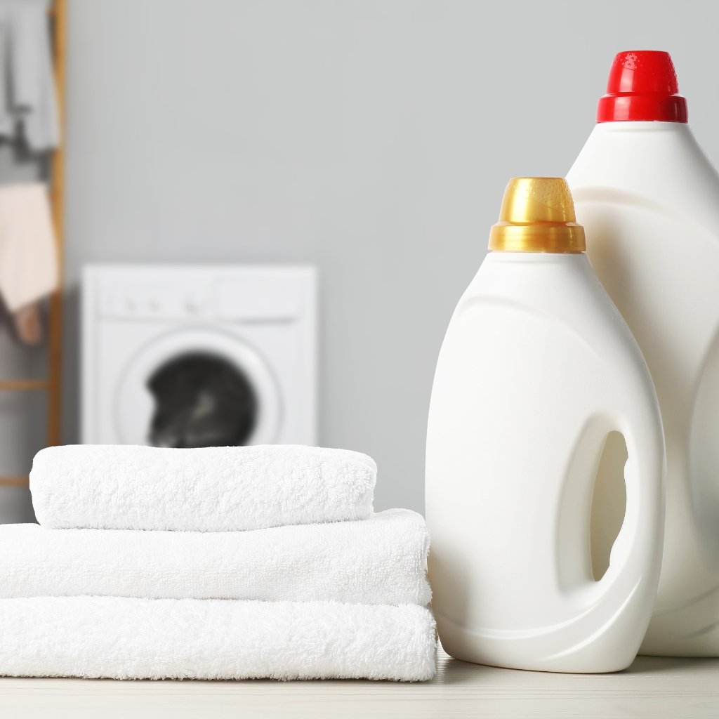 A laundry setup with two white laundry detergent bottles, a neatly folded stack of white towels, and a washing machine in a bright, clean laundry room.

