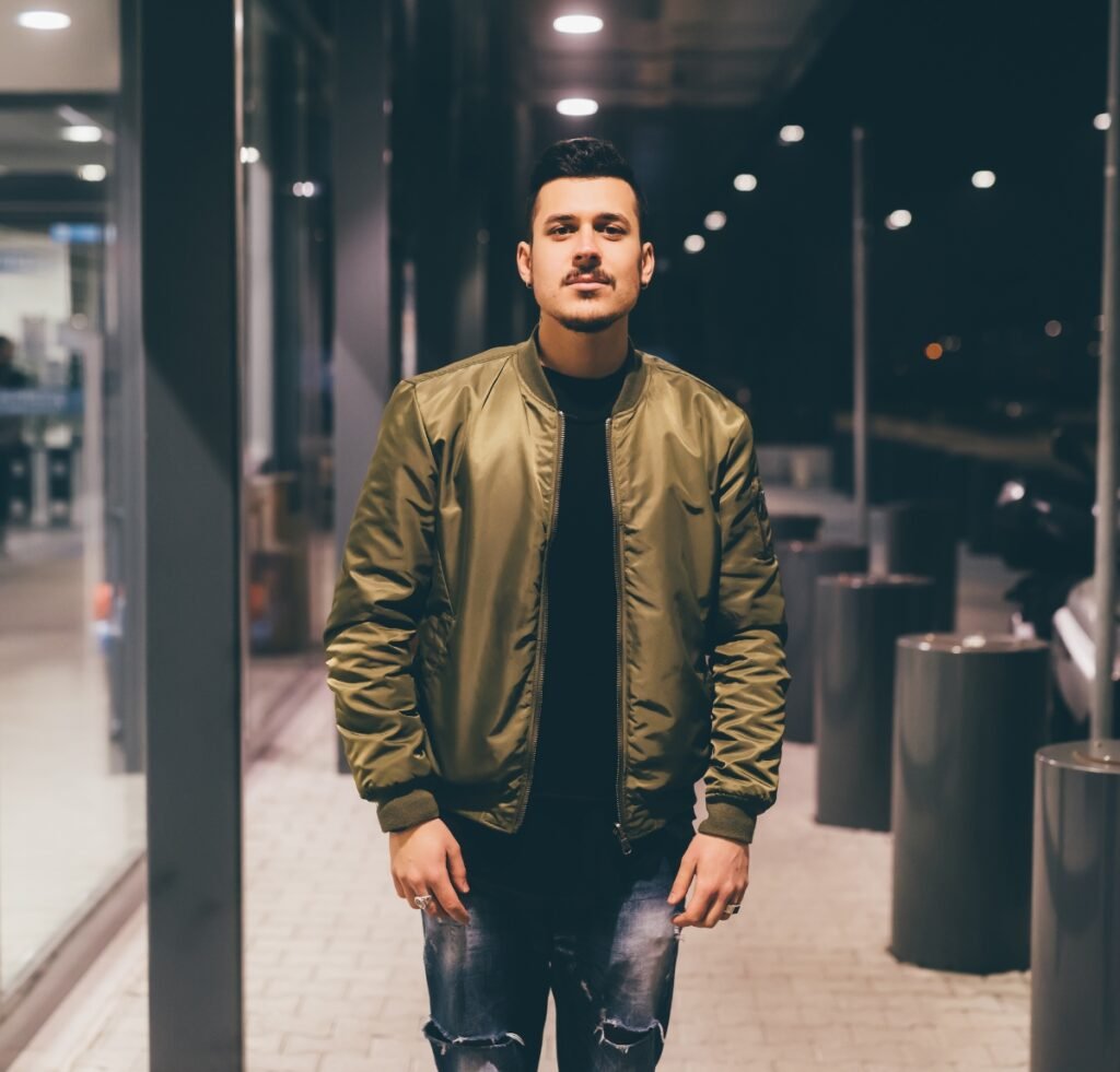 A stylish man stands outdoors at night wearing a olive nylon bomber jacket over a black shirt, paired with distressed jeans.