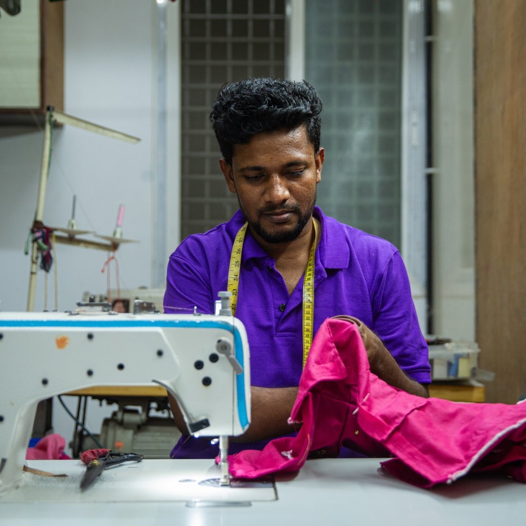 A man in a purple shirt sits at a sewing machine, measuring with a yellow tape as he works on pink fabric in a workshop.
