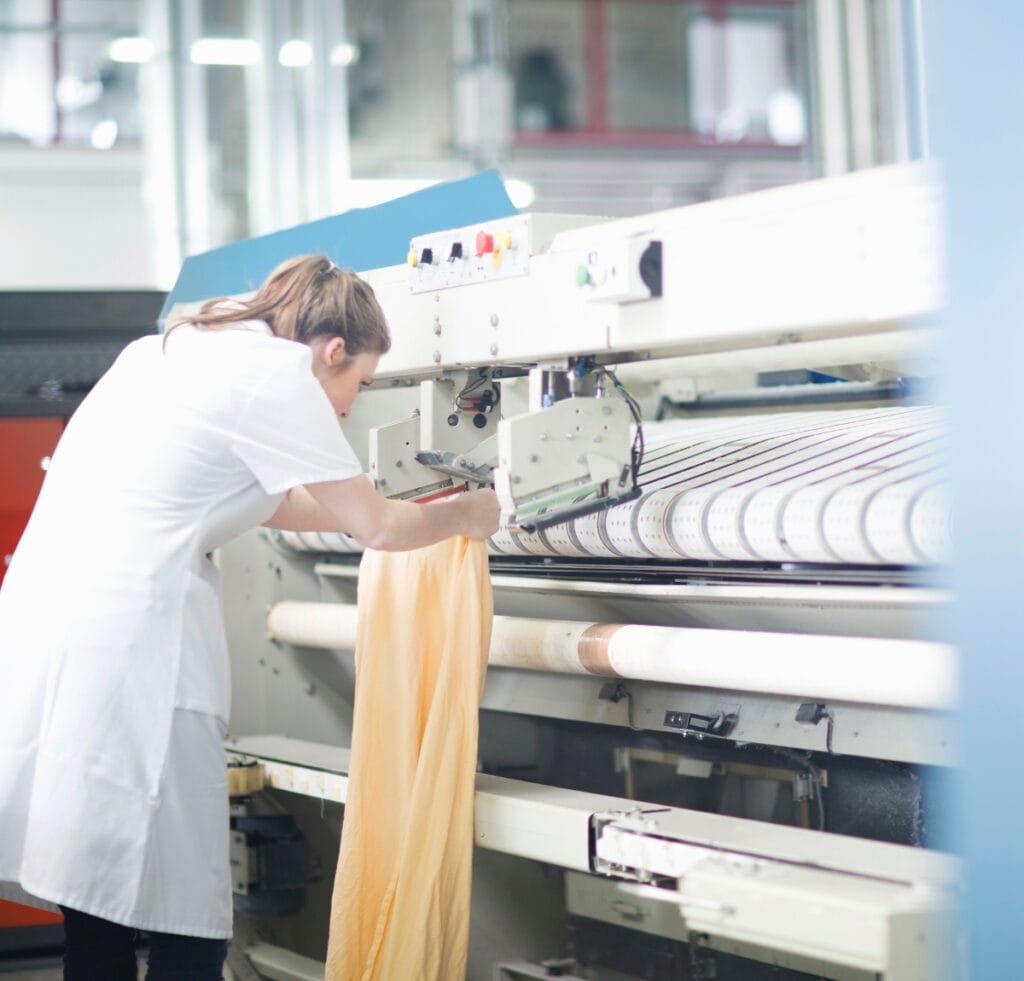 A textile worker in a white coat operates a large weaving/finishing machine, handling a long piece of pale linen fabric fed through the machinery.