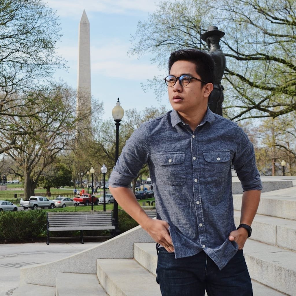A man wearing a chambray cotton shirt stands on steps in a park with the Washington Monument in the background. He has glasses and a watch, hands in pockets, looking to the side.
