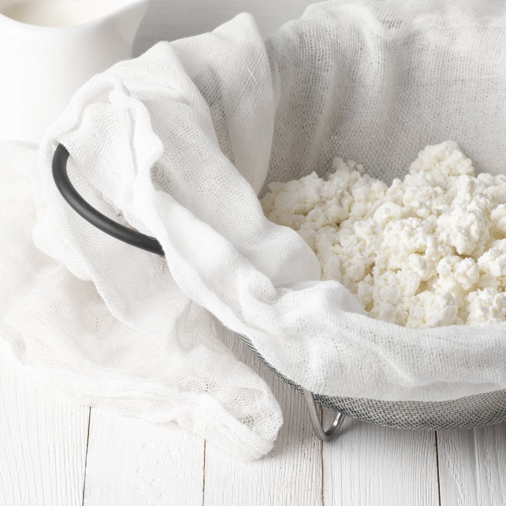 A metal colander lined with a white cheesecloth sits on a white wooden surface, filled with fresh white cottage cheese.