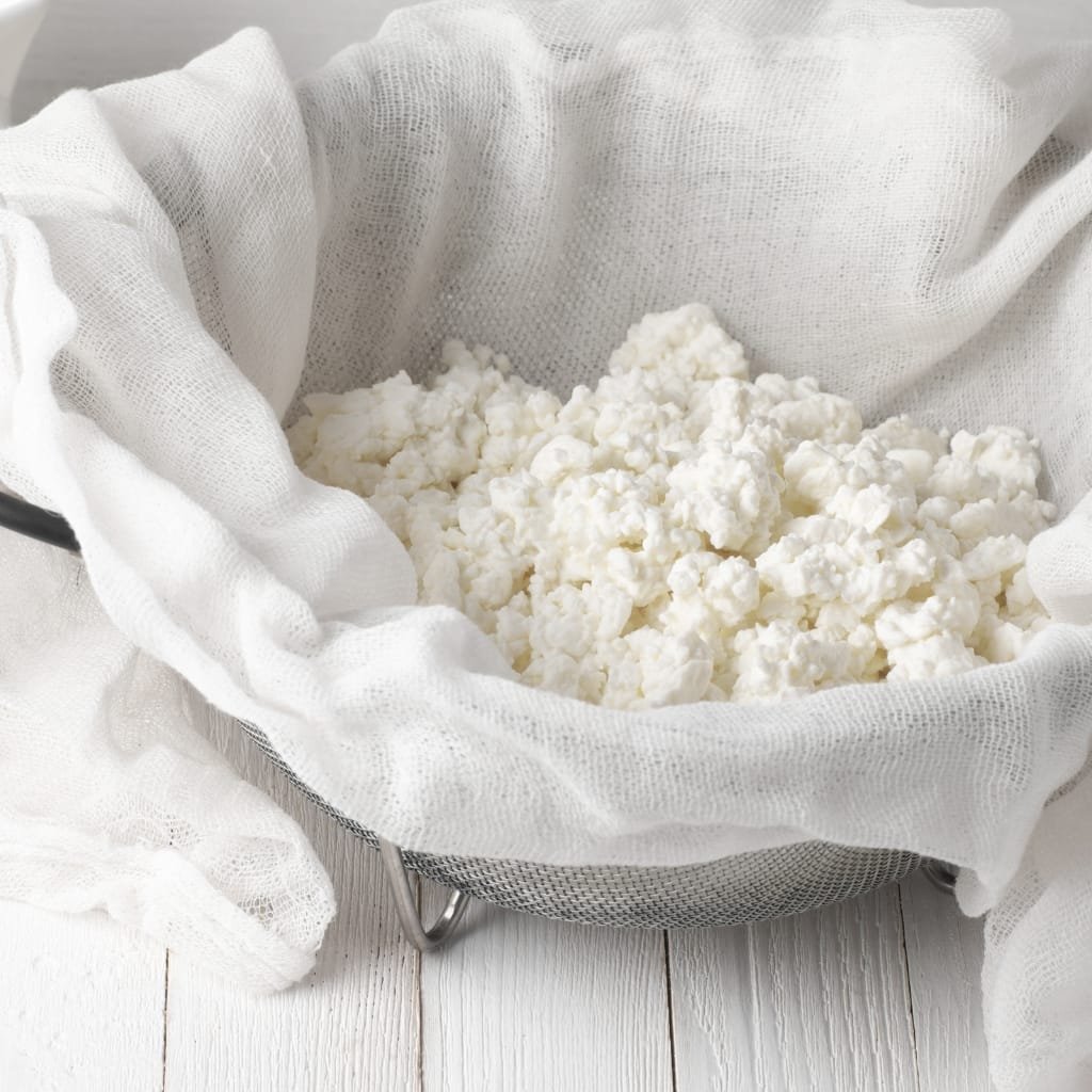 A metal sieve filled with white cottage cheese-like curds, lined with a soft white cheesecloth draped over the sides.