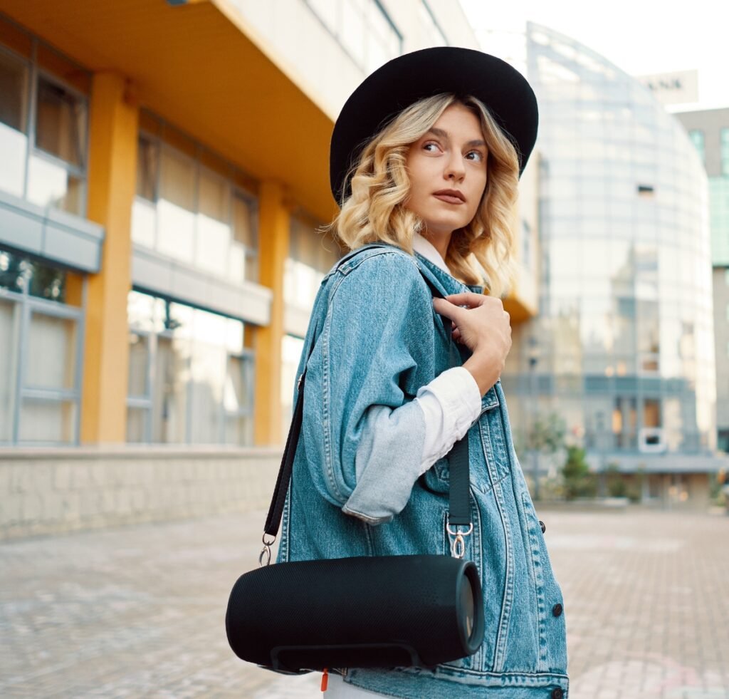 A stylish woman wearing a light-wash denim bomber jacket and a white shirt, paired with a wide-brim black hat. She carries a cylindrical black statement bag over her shoulder, standing outdoors in an urban setting with modern buildings in the background.