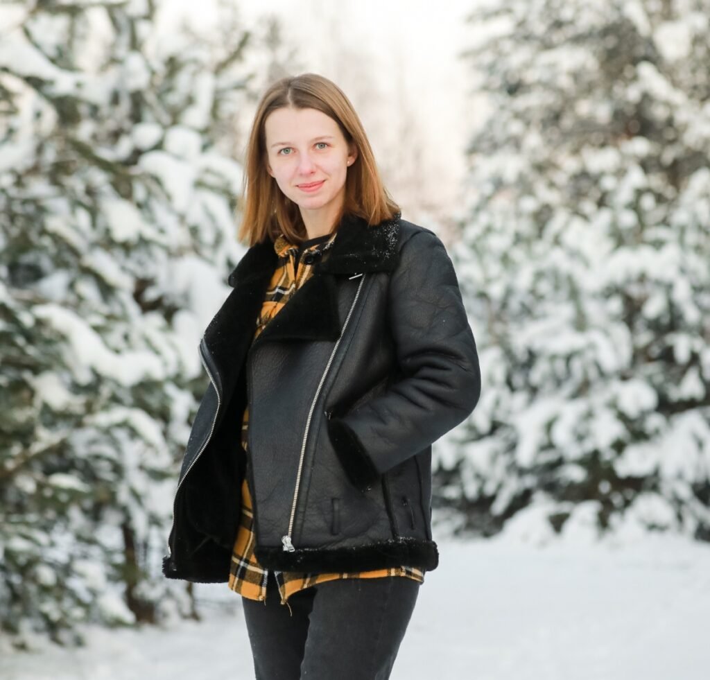 A woman standing outdoors in a snowy scene, wearing a black shearling bomber jacket over a mustard-yellow plaid shirt. Snow-covered trees frame the background.
