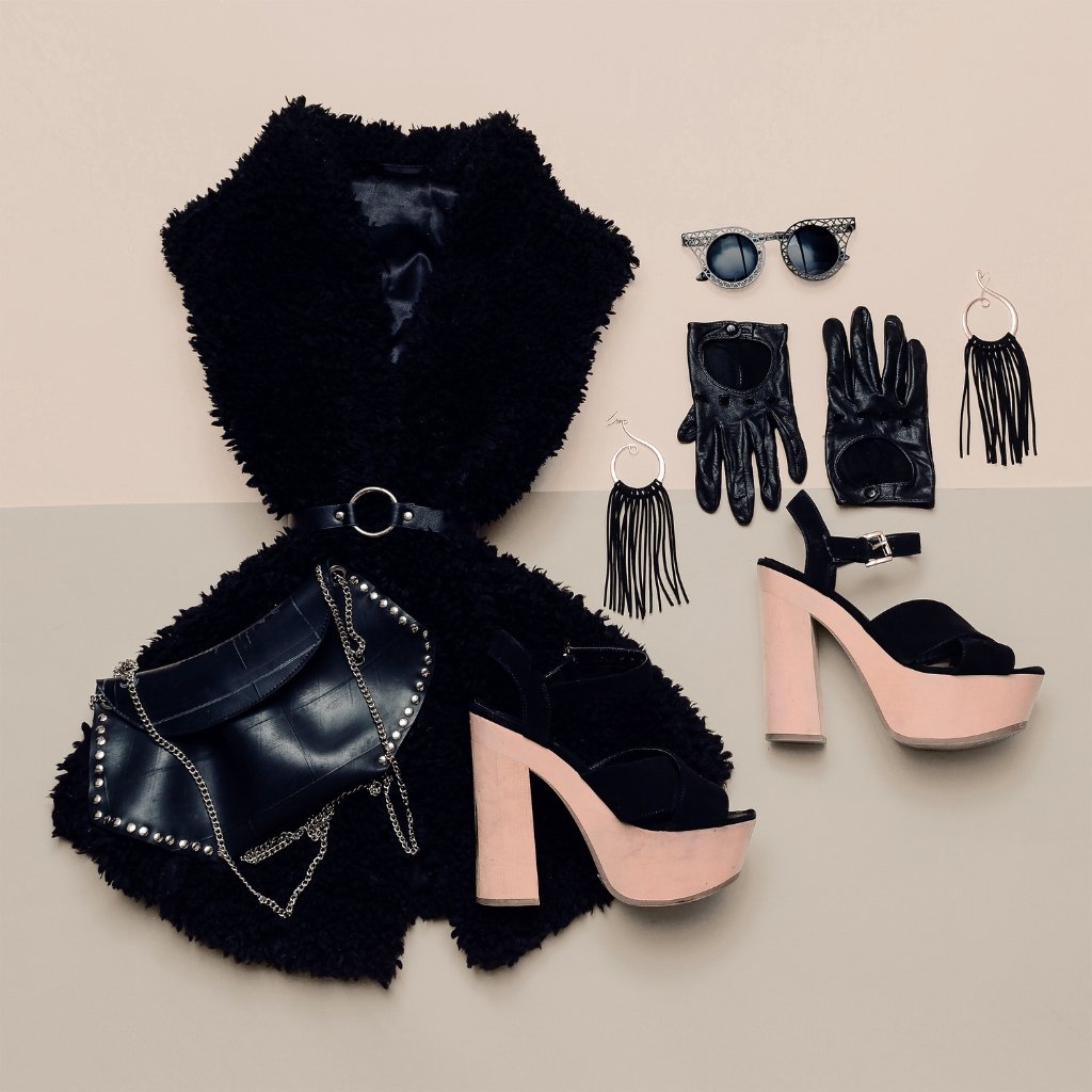 Fashion flat lay featuring a black faux fur vest with a belt, a black quilted bag with silver chains, blush pink platform sandals, and bold accessories (sunglasses, gloves, fringe earrings) arranged on a two-tone background.