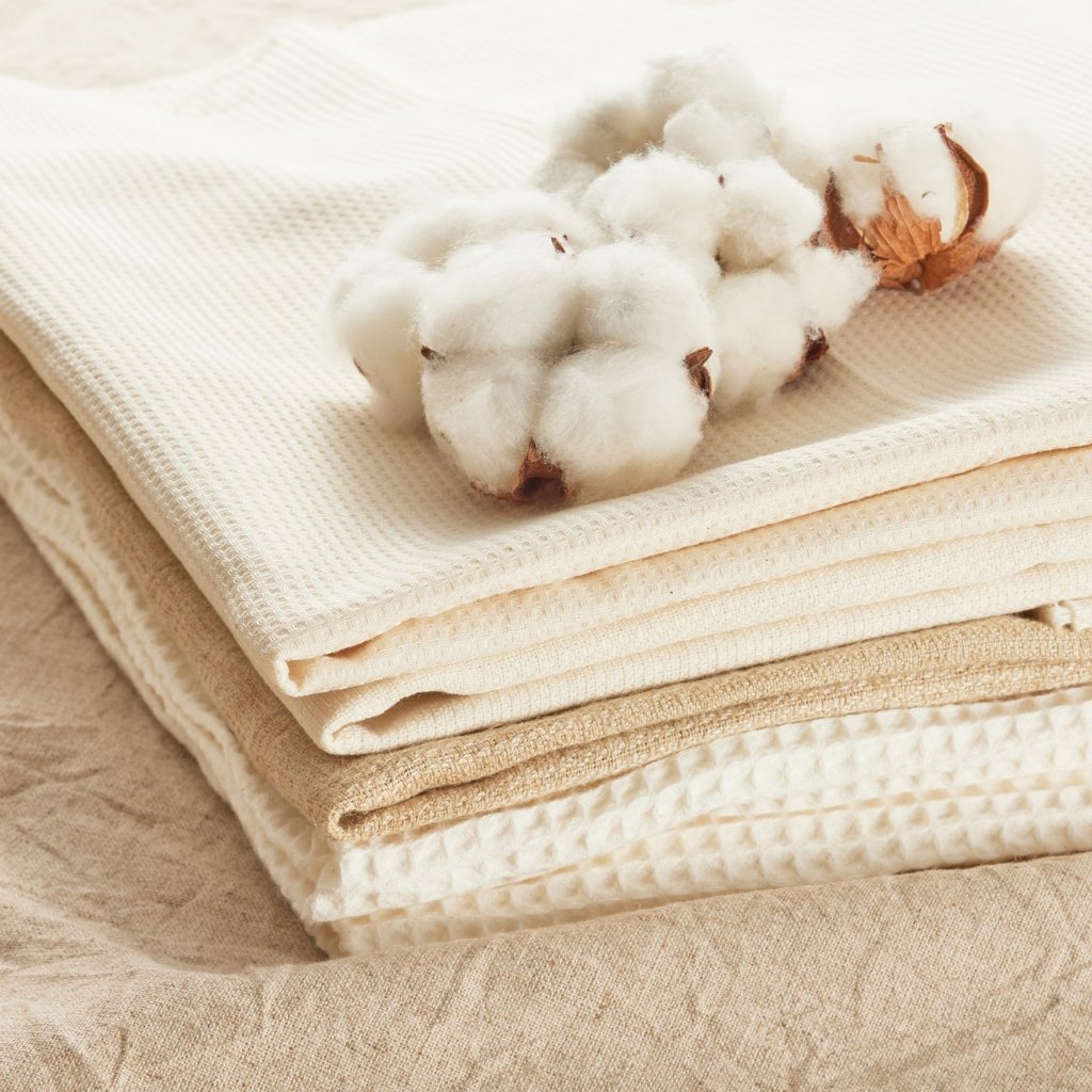 A neatly stacked pile of soft, cream-colored fabrics in varying textures (cotton voile, waffle knit, and jersey) with a small cotton boll resting on top.

