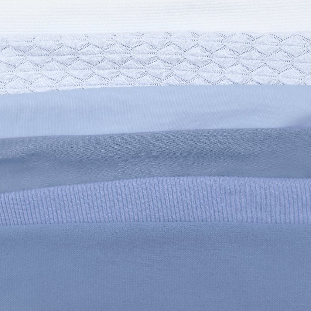 A close-up image of layered fabrics in varying shades of light blue and white. The topmost layer shows a delicate white knit pattern, followed by soft, semi-sheer cotton voile, and several smooth blue fabrics with fine weaves and ribbed edges.