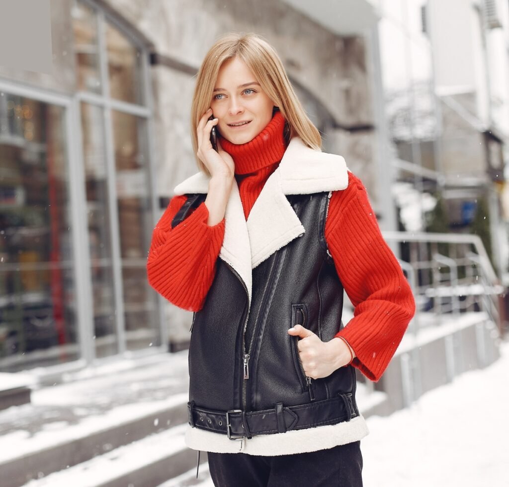 A stylish woman stands outdoors in a snowy urban setting, wearing a black shearling bomber jacket with a white shearling collar and cuffs over a bright red turtleneck sweater. She smiles while talking on her phone, with one hand in a zipper pocket and the other holding the phone.