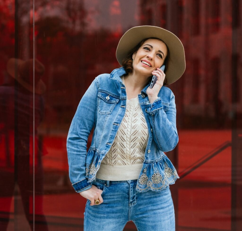 A stylish woman wearing a light denim bomber with lace trim, paired with a cream knit sweater and wide-brim hat, stands outdoors while talking on her phone.
