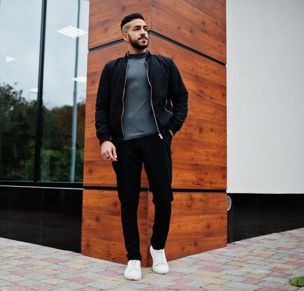 A stylish man stands outdoors beside a modern wooden panel wall, wearing a black slim-fit bomber jacket over a gray sweater, black joggers, and white sneakers. He has a neatly groomed beard and short hair, looking off to the side with one hand in his pocket.