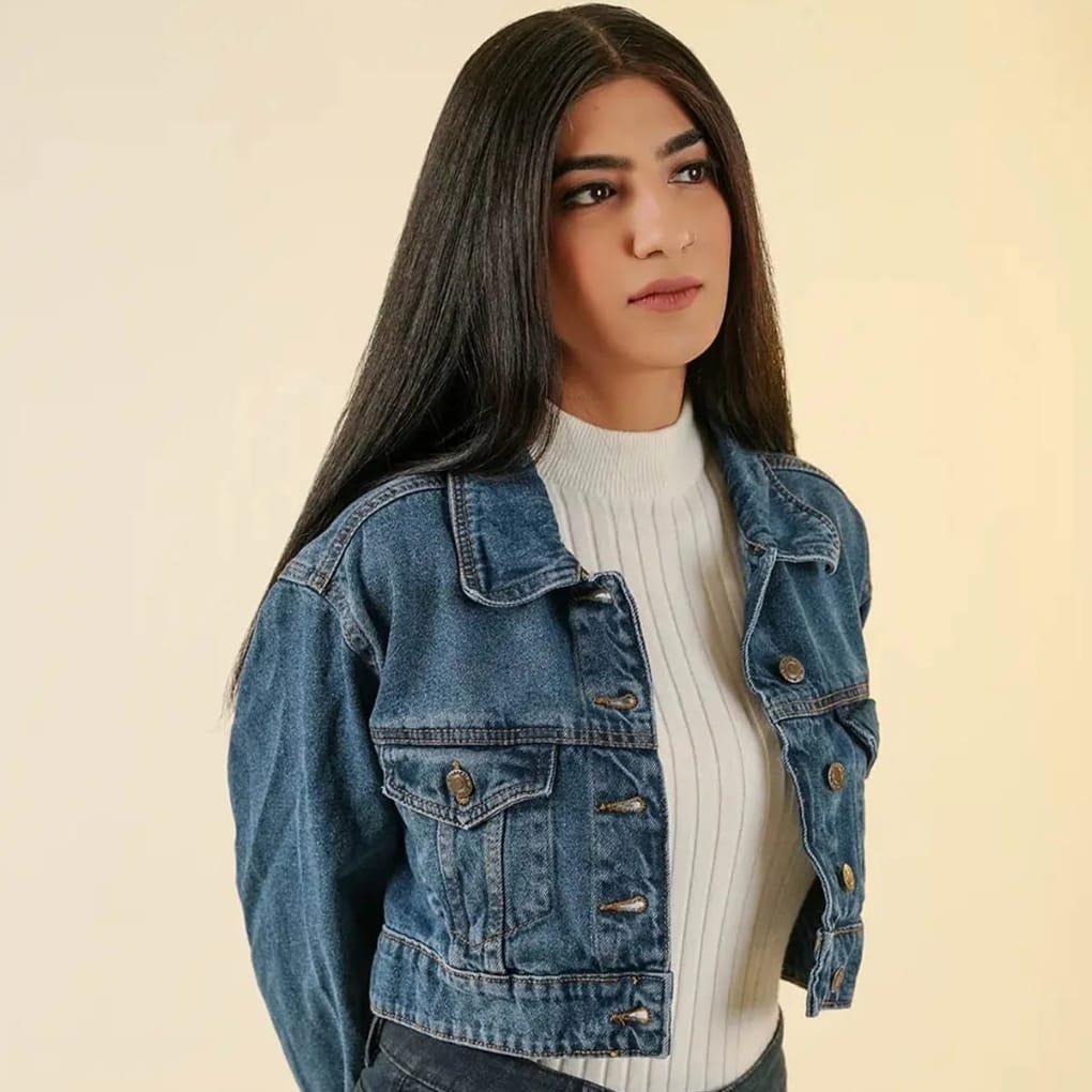 A young woman with long dark hair wearing a light-colored ribbed turtleneck under a blue denim jacket, standing against a pale yellow background.