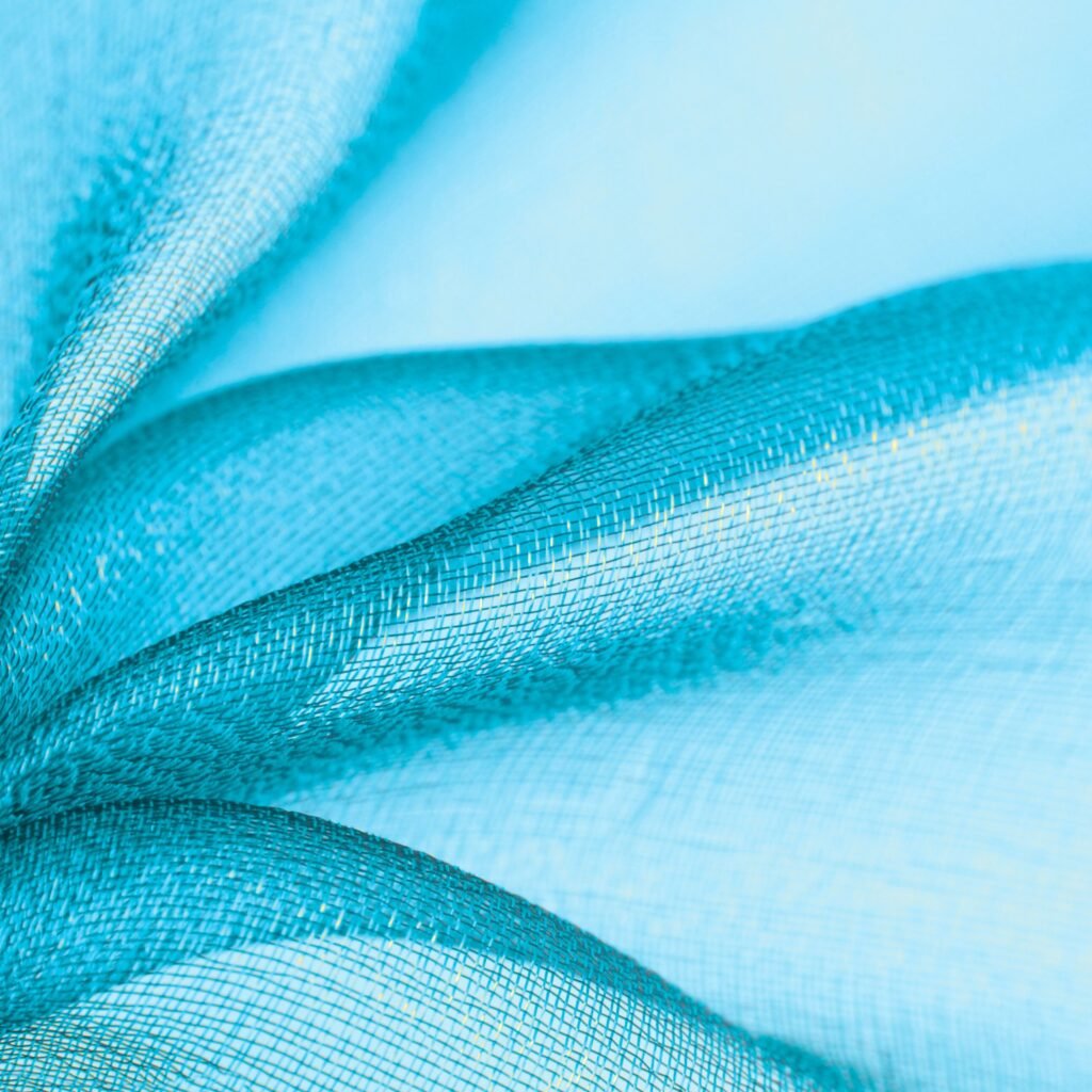 Close-up view of crystal organza fabric: light blue, sheer, delicate mesh with fine weaves and subtle sparkle, softly folded to reveal texture and transparency.