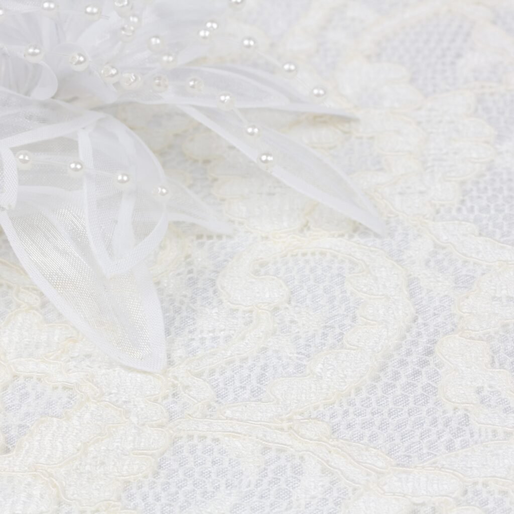 A close-up view of crystal organza fabric featuring delicate sheer ribbons and tiny pearl embellishments arranged on intricate lace, creating a soft, elegant wedding look.