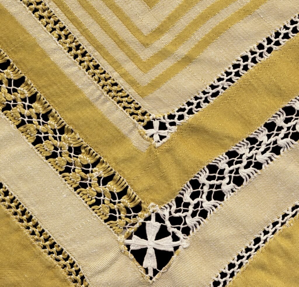 A close-up view of gold and ivory damask linen with geometric patterns and delicate open-work lace trim, arranged in diagonal chevron layers.