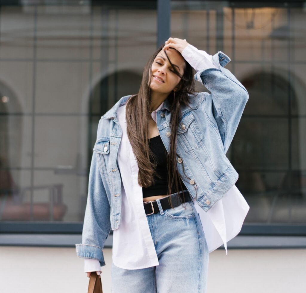 A stylish woman stands outdoors wearing a light-wash denim bomber jacket layered over a black crop top and a white shirt. She pairs it with high-waisted light-wash jeans and a black belt, posing with one hand running through her hair and the other holding a brown bag. The look emphasizes a defined waist and casual-chic vibe.