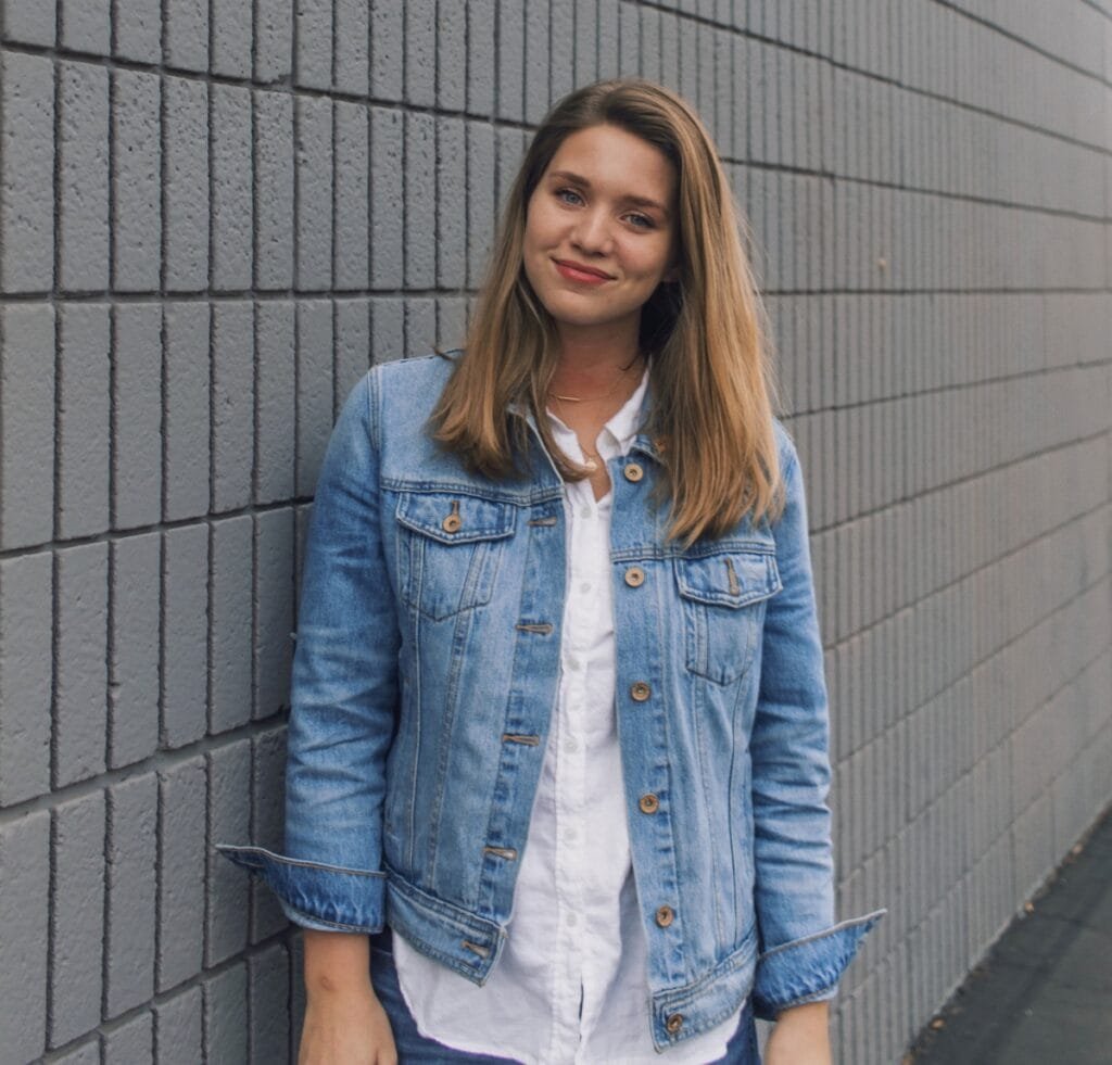 A woman wearing a light-wash denim bomber jacket over a white shirt, standing against a gray brick wall outdoors.